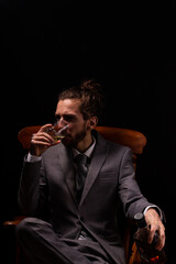A young businessman in a smart gray suit sitting in a wooden arm chair with moody and atmospheric lighting, he is drinking a single shot of whiskey
