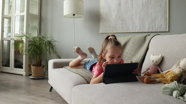 Smiling little girl lying on sofa playing online games, web surfing information, using funny applications on tablet.