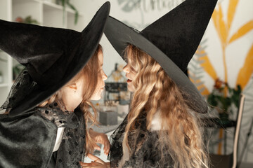 two girls in witch costumes at the Halloween party. Children are wearing black hats. They whisper