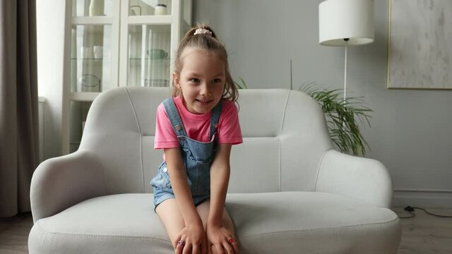Five years little girl with pretty face smiling looking at camera at home sitting on chair near sofa, talking to webcam making online video call or recording vlog, waving hand.