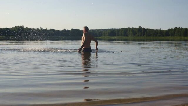 Happy man playing and making splashes standing to belly in lake or river water in summer. Slow motion.