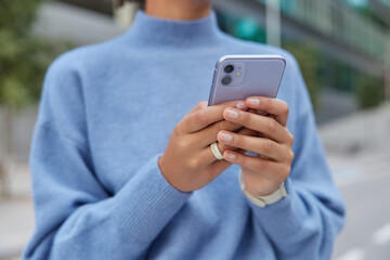 Cropped image of unrecognizable faceless woman in casual blue jumper holds mobile phone types sms message makes online shopping communicates in social networks poses outside uses free internet