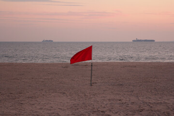 warning red flag on the beach of the Atlantic ocean with two ships on the horizon at sunset 