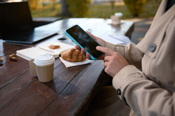 hands of an unrecognizable person wearing beige trench and using a mobile phone on the background of a wooden table with open notepad and copybook and delicious breakfast