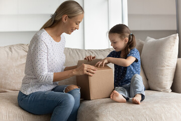 Fototapeta na wymiar Happy young mother unpacking cardboard box with interested cute little preschool kid daughter, feeling excited of getting parcel with wished purchase item from internet store, online shopping concept.