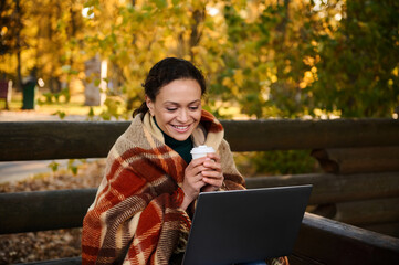 Smiling middle aged woman wrapped in warm checkered blanket holds coffee paper cup, studying ,...