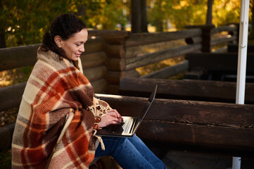 Self-confident middle aged woman wrapped in warm checkered blanket typing on laptop, studying ,...