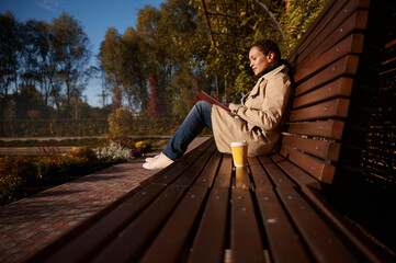 Beautiful peaceful young brunette sitting on wooden bench in park, reading book and drinking...