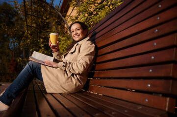 Attractive woman in casual clothing resting on her day off with a book and a takeaway paper glass...