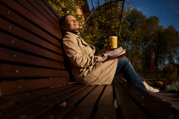 Attractive woman with closed eyes sits on wooden bench in park, holding paper cup of hot drink and...