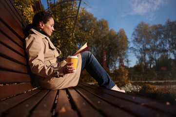 Fototapeta premium Relaxed pretty woman in casual denim and beige coat with a book and a takeaway paper cup of hot drink enjoying a warm sunny autumn weekend in country park, away from the hustle and bustle of the city