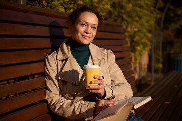 Beautiful serene woman holding a cardboard cup with a hot drink and reading a book while resting on...
