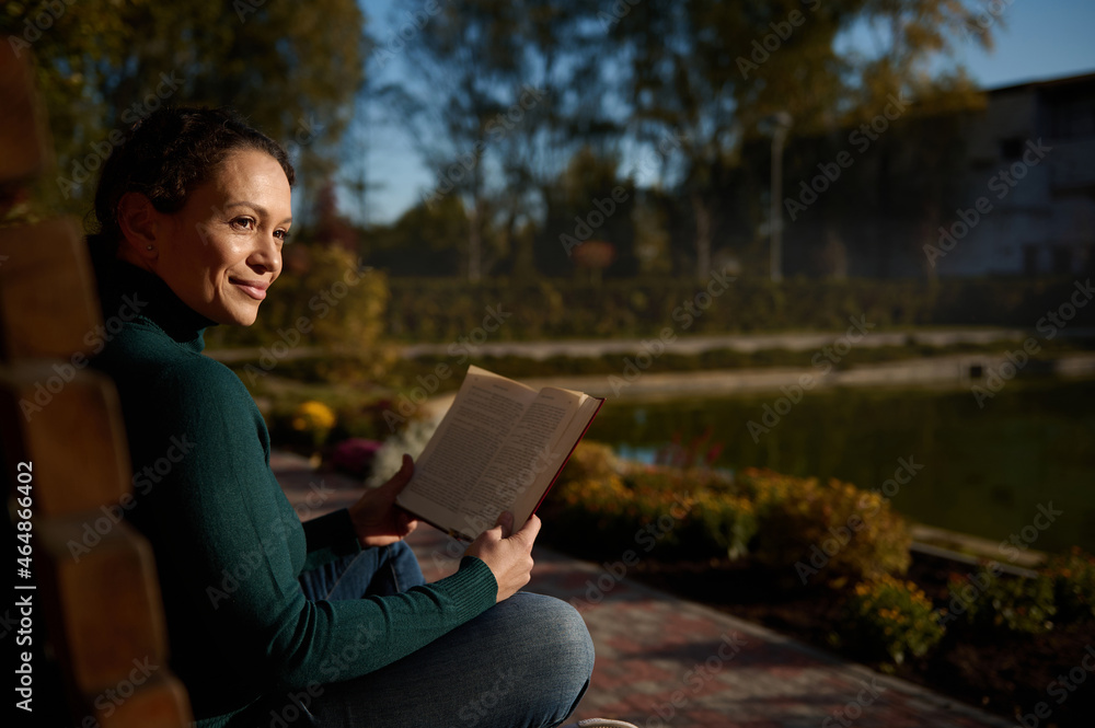 Wall mural Portrait of a contented pretty woman sitting on wooden bench in park, reading book, enjoying warm sunny autumn day , away from the hustle and bustle of city, resting from digital gadgets and work