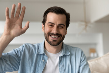 Happy millennial handsome man waving hands looking at camera, starting distant conversation. Smiling friendly young male blogger recording video or steaming stories online, siting on sofa at home.