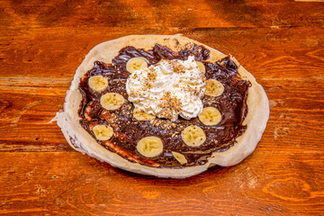 Open chocolate crepe with banana, whipped cream and nuts on wooden table