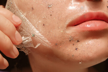 Fototapeta premium Young woman with healthy white pale skin removes purifying transparent peel off mask with shiny sparkling star glitter from her face. Trendy cosmetic beauty procedure.Skin care concept.