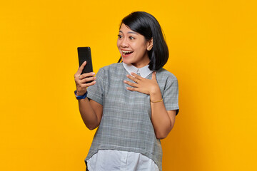 Cheerful young Asian woman looking at mobile phone and keeping palms on chest over yellow background