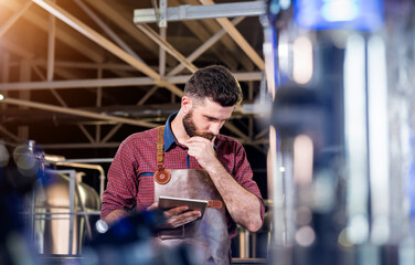 Young male brewer in leather apron supervising the process of beer fermentation at modern brewery...