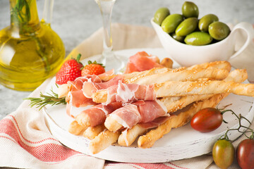 Traditional Italian snacks for wine, breadsticks (grissini), tomatoes, prosciutto (jamon), strawberries and olives. Selective focus.