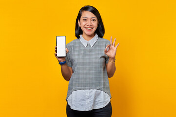 Cheerful Asian woman showing smartphone blank screen and gesturing okay sign over yellow background