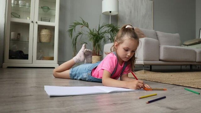 Smilling happy girl lying on warm floor with a toy elephant near to her enjoying creative activity, drawing pencils coloring pictures in albums.