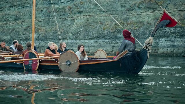 Vikings Sail on an Old Ship with a Lowered Sail on a Quiet River Against the Backdrop of a Rocky Coast. The Men Row the Oars Diligently Towards Adventure. Medieval Reconstruction.