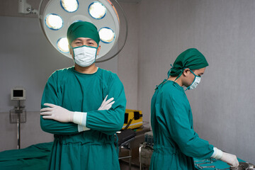 Fototapeta na wymiar Doctors in operating room wearing green mask surgical gown surgeon hair cap latex glove one stand with arms crossed in front of bed and electrical defibrillator other while doctor prepare instruments