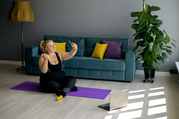 An obese young blonde woman with a dumbbell takes a selfie on her smartphone during a home workout. Sports at home in the living room with a green sofa and yellow-purple pillows. Model plus size