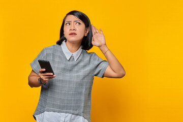 Portrait of serious looking asian woman trying to overhear secret conversation and holding smartphone over yellow background