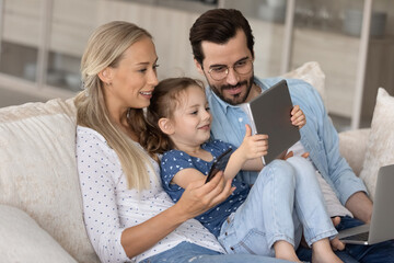 Obraz premium Addicted to modern technology happy young family couple with adorable little preschool child daughter using different gadgets, playing mobile games online or watching cartoons, spending time online.