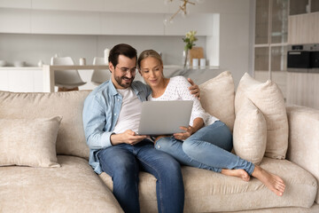 Fototapeta premium Happy loving young family couple looking at laptop screen, sitting on cozy sofa at home, enjoying watching comedian movie, web surfing information or spending time online in social network on weekend.