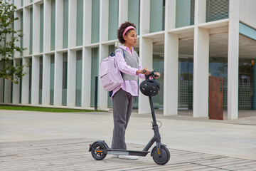 Female student rides to university by electric scooter carries rucksack poses against city...