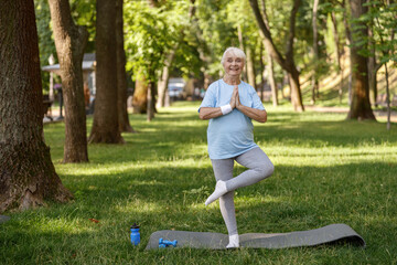 Happy senior lady in tracksuit does tree pose practicing yoga asana in park