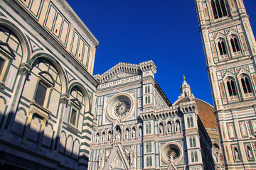 Naklejka premium The elaborated facade of the Cathedral of Santa Maria del Fiore in Florence, Firenze, Italy, with the Bell Tower and detail of the Dome and deep blue sky in background.