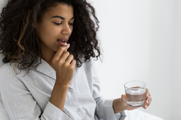 Young afro american woman hold glass of water, take pill