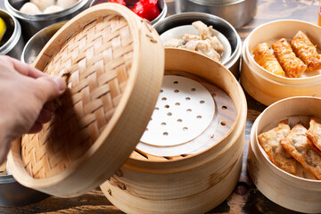 A view of a hand opening the lid to the top of a stack of bamboo steamer containers, among a variety of Chinese dumplings.