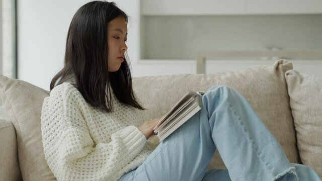 Happy Asian woman in a white sweater sitting on the couch and reading a book at home