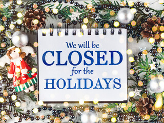 Signboard with the inscription We will be closed for the holidays and green branches of a Christmas tree. View from above, closeup, indoors, no people. Congratulations for family, friends, colleagues