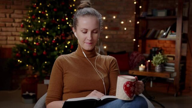Woman at home in December, Winter. Reading book listening music and drinking hot tea, Christmas decoration in cozy dark room.