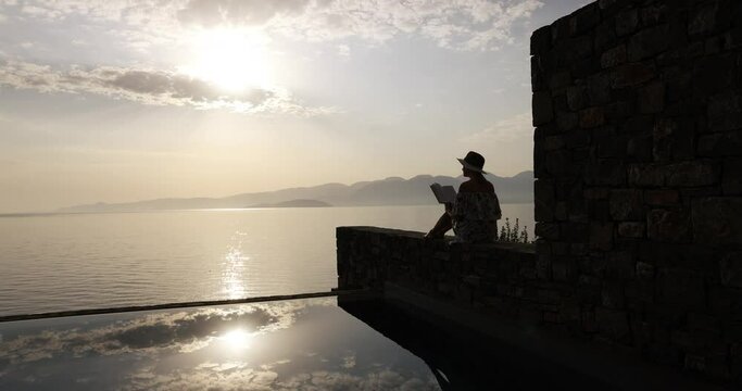 relaxing vacation - woman reading a book near swimming pool with sea view in luxury resort at sunrise