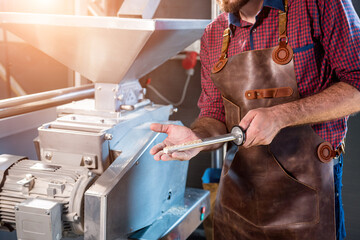 A young brewer in a leather apron controls the grinding of malt seeds in a mill at a modern brewery - obrazy, fototapety, plakaty