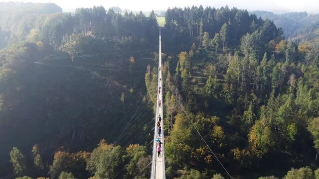 Video from a drone flying over a suspended wooden bridge with steel ropes over a dense forest in West Germany, visible tourists walking on the bridge.