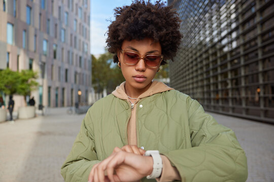Serious stylish curly haired young woman looks at watch checks time waits for somone wears sunglasses casual hoodie and jacket poses at street among cityscrapers during daytime comes on meeting