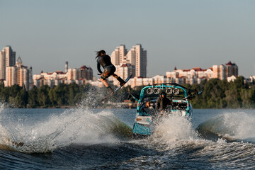 wakeboarder moving fast behind boat holding rope and jumping high on splashing river wave.