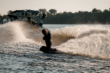 Athletic guy holds rope and balancing on wakeboard behind motor boat on splashing river waves.