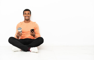 African American man sitting on the floor over isolated copyspace background holding coffee to take away and a mobile