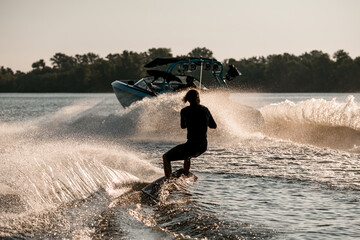 Rear view of active man riding wakeboard behind motor boat on splashing river waves. Active and extreme sports - obrazy, fototapety, plakaty