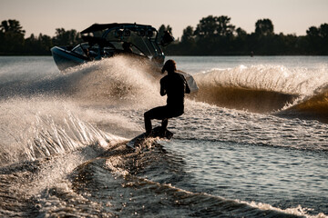 Rear view of energy man riding wakeboard behind motor boat on splashing river waves. Active and...
