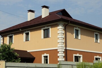 big private house with windows under a brown tiled roof with a chimneys behind a gray fence against the blue sky