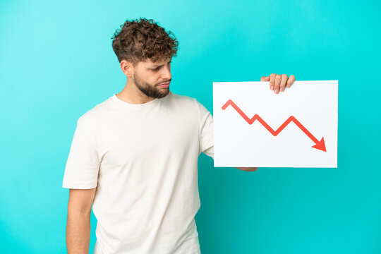 Young handsome caucasian man isolated on blue background holding a sign with a decreasing statistics arrow symbol with sad expression